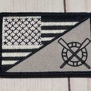 May include: Embroidered patch featuring a split design. One half displays a black and white American flag, the other a gray background with a life preserver and crossed oars. The patch has a black border.