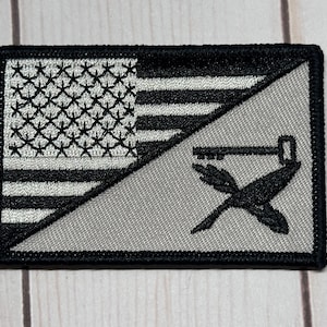 May include: A rectangular patch featuring a split design. The top left corner displays a black and white American flag. The lower right section has a gray background with a black key, hammer, and crossed arrows design. The patch is bordered in black.