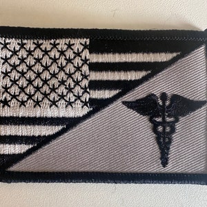 May include: A gray and black embroidered patch with an American flag on the left side and a medical symbol on the right side.