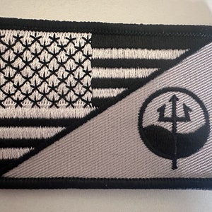 May include: A black and white embroidered patch with a US flag on one side and a trident logo on the other. The patch is sewn onto a gray fabric background.