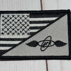 May include: Embroidered patch featuring a split design. The left side displays the American flag in black and white, while the right side showcases a gray background with a black emblem of wings and a symbol. The patch has a black border.