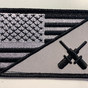 May include: A gray and black embroidered patch with an American flag on the left side and a pair of crossed screwdrivers on the right side.