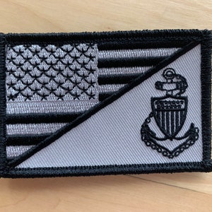 May include: A black and white embroidered patch with a US flag on one side and a Coast Guard anchor on the other side.