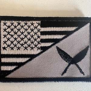 May include: A black and white embroidered patch with a diagonal split. The top half features a stylized American flag with white stars and red stripes. The bottom half is gray with a black stylized symbol of two crossed feathers.