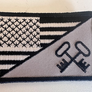 May include: A gray and black embroidered patch with a diagonal split. The top half features a stylized American flag with white stars on a black background. The bottom half features two crossed keys in black on a gray background.