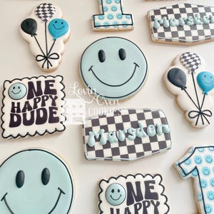 May include: A collection of decorated sugar cookies featuring light blue and black color schemes. Shapes include the number one, smiley faces, balloons, and rectangular plaques with the name Marsden and the text One Happy Dude.