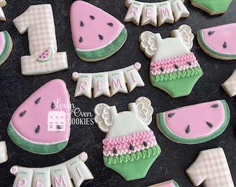 Puede incluir: Surtido de galletas de azúcar decoradas, que incluyen rebanadas de sandía, galletas con el número "1" y galletas con forma de body de bebé. Las galletas son rosas, verdes y blancas, con detalles en negro. Algunas galletas deletrean el nombre "REMI".