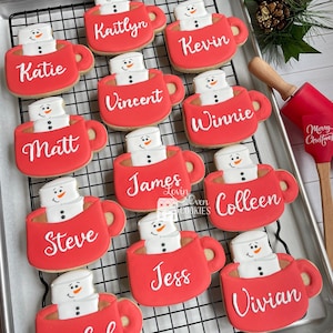 Puede incluir: Galletas navideñas con forma de tazas rojas con muñecos de nieve de malvavisco. Cada galleta tiene un nombre como "Katie" o "Kevin". Las galletas están sobre una rejilla, con decoraciones festivas en el fondo.