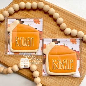 Puede incluir: Dos galletas con forma de porción de tarta de calabaza decoradas con glaseado naranja y bordes de masa blanca. Una galleta tiene el nombre Rowan y la otra Isabelle en letras blancas. Cada galleta está en una caja cuadrada transparente.