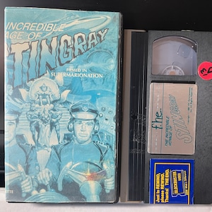 May include: A VHS tape of the "The Incredible Voyage of Stingray" with a blue and silver label. The label has the text "The Incredible Voyage of Stingray" and "fhe." The tape is in a black case with a red sticker that says "13".