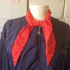 May include: A red and black polka dot scarf tied around the neck of a navy blue jacket. The scarf is tied in a simple knot and the ends are hanging down. The jacket has a zipper and a silver zipper pull with a deer design.