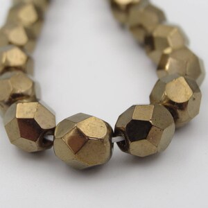 May include: A string of faceted, metallic gold beads. Each bead has a geometric, multi-sided shape. The beads are strung on a white string, and the background is plain white. These beads are suitable for jewellery making.