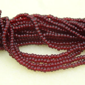 Könnte beinhalten: Nahaufnahme von Strängen kleiner, runder, dunkelroter Perlen. Die Perlen sind aufgefädelt und erzeugen einen strukturierten, geschichteten Effekt. Die Perlen haben eine tiefe, durchscheinende rote Farbe, und der Gesamteffekt ist einer von Reichtum und Tiefe.