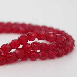 May include: Strands of faceted, deep red beads. The beads have a geometric shape and are strung together, creating a flowing, curved line. The beads are a rich, saturated colour, and the image is set against a plain white background.