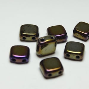 Puede incluir: Primer plano de varias cuentas cuadradas con dos agujeros cada una. Las cuentas tienen un acabado metálico con colores que van del dorado al morado. Están dispersas sobre una superficie blanca.