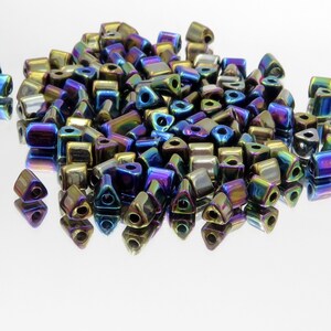 May include: A collection of iridescent, geometric beads in various shapes, including squares and triangles. The beads have a metallic finish with colors shifting from gold to blue and purple. They are scattered on a reflective surface.