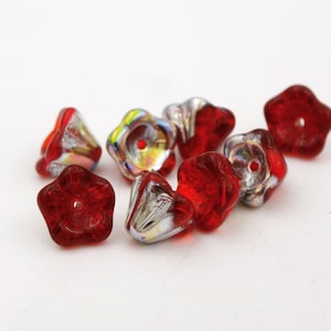 Puede incluir: Un conjunto de cuentas de vidrio rojas en forma de flor con un revestimiento plateado iridiscente. Las cuentas tienen un agujero en el centro y están dispersas sobre una superficie blanca. Estas cuentas son adecuadas para la fabricación de joyas y manualidades.