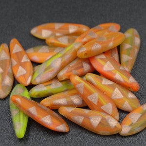 May include: A collection of orange, yellow, and green glass beads. Each bead is elongated with a pointed end and decorated with a copper-colored triangle pattern. The beads are scattered on a dark surface.