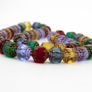 May include: A strand of faceted beads in various colors, including green, purple, yellow, and red. The beads have a textured, multi-faceted surface and a gold-colored interior. The beads are strung together, creating a curved shape against a white background.