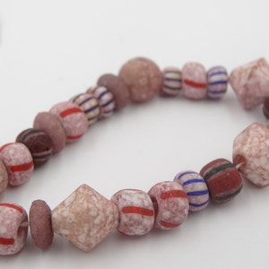 May include: A string of mixed beads in pink, red, and brown hues. The beads are of various shapes, including round, cylindrical, and faceted, some with stripes or speckled designs. The beads are strung on a white string.