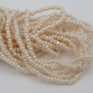May include: Close-up of multiple strands of small, off-white, pearl-like beads. The beads are strung together, creating a textured and layered effect. The beads have a subtle luster and are arranged against a plain white background.