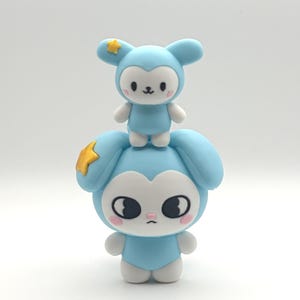May include: Two stacked light blue and white animal figurines with large ears. The top figure has a small yellow star on its ear, while the larger bottom figure features a yellow star on its side. Both have white faces, pink cheeks, and black eyes.