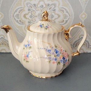 May include: Cream ceramic teapot with gold detailing and a matching lid. The teapot has a floral pattern in blue and pink, with a gold handle and spout. A classic design for serving tea, ideal for a tea party.