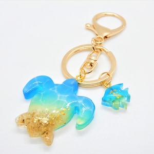Peut inclure: Un porte-clés en or avec un charme de tortue en résine bleue et verte et un plus petit charme de poisson bleu et vert. Le charme de la tortue a un effet de sable doré scintillant.