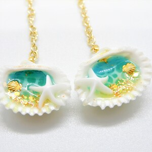 K&ouml;nnte beinhalten: Ein Paar Ohrringe mit goldener Kette und einem wei&szlig;en Muschel-Anh&auml;nger. Jede Muschel enth&auml;lt blaues Harz mit goldenem Glitzer, einen wei&szlig;en Seestern und eine kleine goldene Muschel.