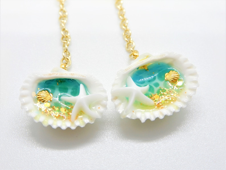 K&ouml;nnte beinhalten: Ein Paar Ohrringe mit goldener Kette und einem wei&szlig;en Muschel-Anh&auml;nger. Jede Muschel enth&auml;lt blaues Harz mit goldenem Glitzer, einen wei&szlig;en Seestern und eine kleine goldene Muschel.