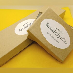 K&ouml;nnte beinhalten: Zwei braune Pappkartons mit wei&szlig;en ovalen Aufklebern, auf denen "Resinfairytales" und "resinfairytales.etsy.com" steht. Die Kartons befinden sich auf einer gelben Oberfl&auml;che.