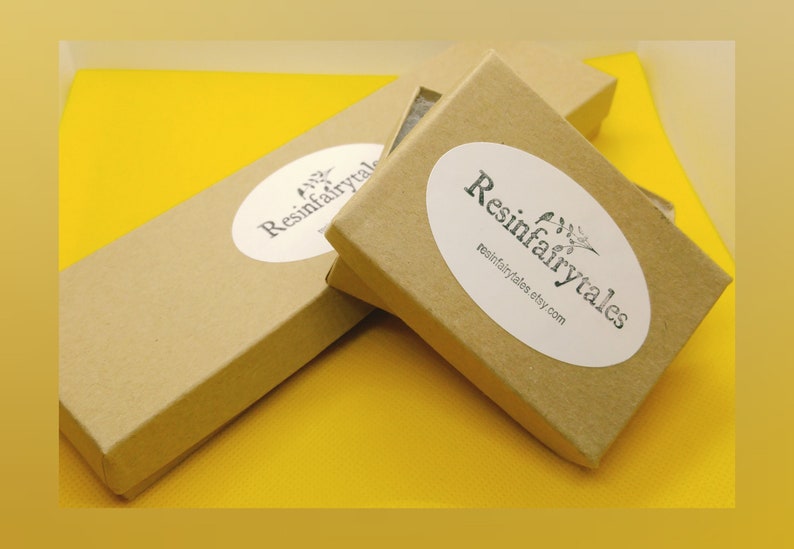 K&ouml;nnte beinhalten: Zwei braune Pappkartons mit wei&szlig;en ovalen Aufklebern, auf denen "Resinfairytales" und "resinfairytales.etsy.com" steht. Die Kartons befinden sich auf einer gelben Oberfl&auml;che.