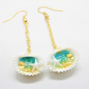 K&ouml;nnte beinhalten: Ein Paar goldene Ohrringe mit einem Muschel-Anh&auml;nger. Die Muscheln sind mit blauem und gr&uuml;nem Harz, goldenem Glitzer und wei&szlig;en Seesternen gef&uuml;llt.