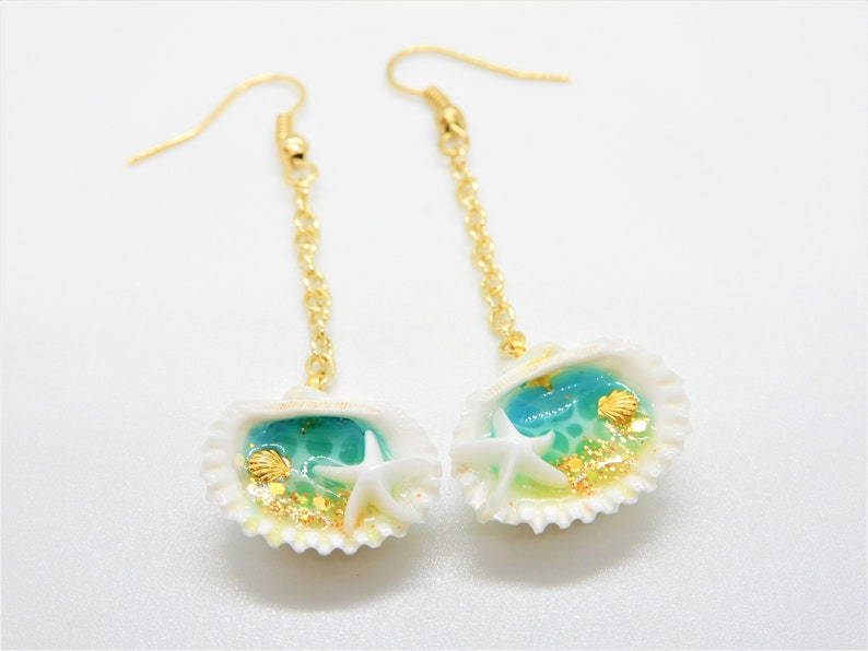 K&ouml;nnte beinhalten: Ein Paar goldene Ohrringe mit einem Muschel-Anh&auml;nger. Die Muscheln sind mit blauem und gr&uuml;nem Harz, goldenem Glitzer und wei&szlig;en Seesternen gef&uuml;llt.