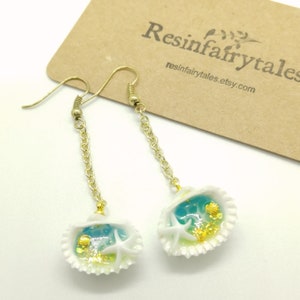 K&ouml;nnte beinhalten: Ein Paar goldene Ohrringe mit einem wei&szlig;en Muschel-Anh&auml;nger. Der Muschel-Anh&auml;nger enth&auml;lt blaues und gr&uuml;nes Harz mit goldenem Glitzer, einen wei&szlig;en Seestern und eine kleine wei&szlig;e Muschel.