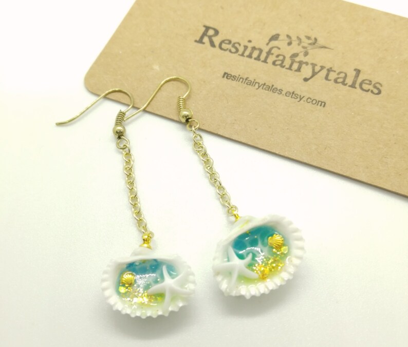 K&ouml;nnte beinhalten: Ein Paar goldene Ohrringe mit einem wei&szlig;en Muschel-Anh&auml;nger. Der Muschel-Anh&auml;nger enth&auml;lt blaues und gr&uuml;nes Harz mit goldenem Glitzer, einen wei&szlig;en Seestern und eine kleine wei&szlig;e Muschel.