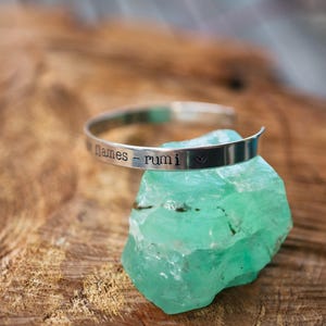 May include: A silver bracelet with the inscription "names - rumi" and a small heart symbol, resting on a light green crystal.