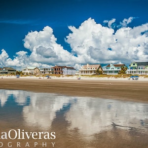 May include: A scenic beach landscape featuring a row of colorful houses along the shoreline under a bright blue sky filled with fluffy white clouds. The wet sand reflects the sky and clouds, creating a mirror effect. The image is a photograph.