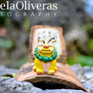 May include: A yellow ceramic figurine of a creature with green beads around its neck. The figurine is sitting on a brown tile.