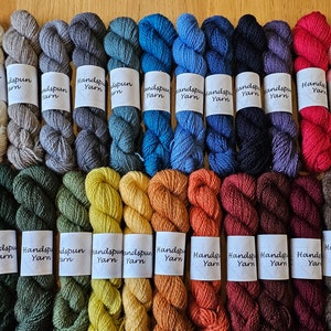 May include: A collection of handspun yarn skeins in a variety of colors, including shades of green, blue, orange, and pink. Each skein has a label that reads "Handspun Yarn". The yarn is arranged on a wooden surface.