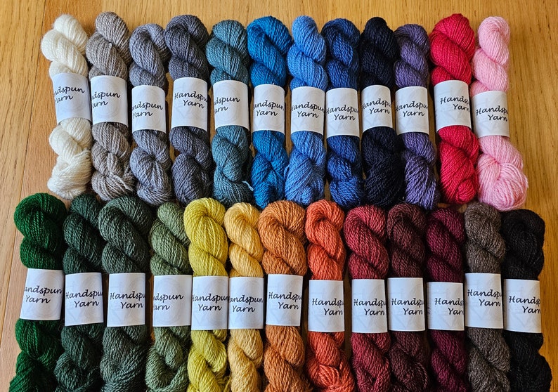 May include: A collection of handspun yarn skeins in a variety of colors, including shades of green, blue, orange, and pink. Each skein has a label that reads "Handspun Yarn". The yarn is arranged on a wooden surface.