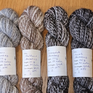 Könnte beinhalten: Vier Knäuel Garn in verschiedenen Farben und Texturen, beschriftet mit der Wollart und dem Gewicht. Die Etiketten geben auch die Länge des Garns in Metern und das Gewicht in Gramm an.