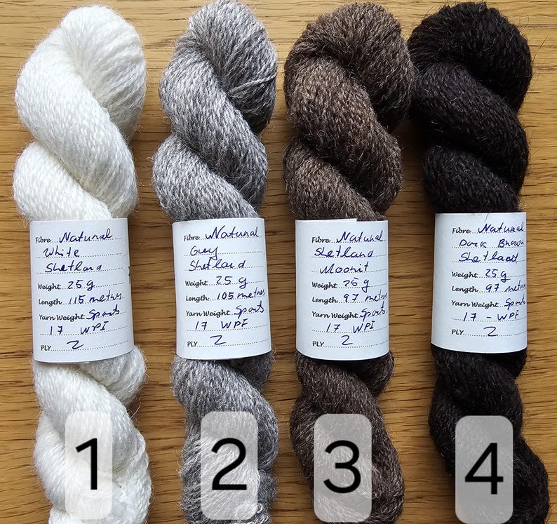 May include: Four skeins of natural Shetland wool yarn in white, gray, brown, and dark brown. Each skein is labeled with fiber details, weight, and length. The yarn is suitable for knitting and other fiber crafts.