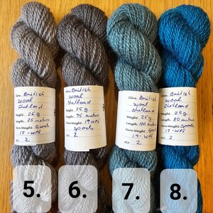 May include: Four skeins of British Shetland wool yarn in shades of gray, teal, and blue. Each skein is labeled with weight, length, and yarn weight details. The yarn is labeled as sports weight.
