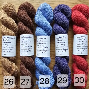 May include: Five skeins of colorful yarn in shades of tan, brown, blue, purple, and red. Each skein has a label with handwritten details. The yarn is arranged on a wooden surface, with the numbers 26, 27, 28, 29, and 30 visible.