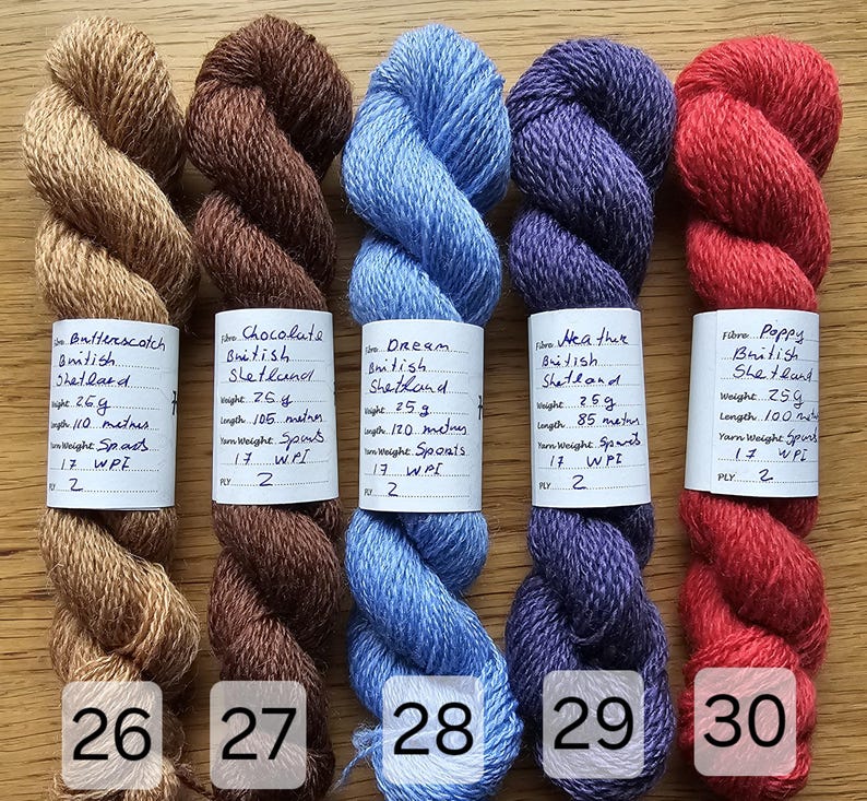 May include: Five skeins of colorful yarn in shades of tan, brown, blue, purple, and red. Each skein has a label with handwritten details. The yarn is arranged on a wooden surface, with the numbers 26, 27, 28, 29, and 30 visible.