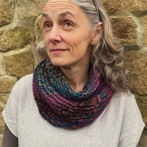 May include: A woman with gray hair wearing a colorful knitted infinity scarf. The scarf is made with a variety of colors, including purple, blue, green, and orange.