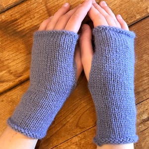 Könnte beinhalten: Ein Paar gestrickte Fäustlinge in einem weichen, blau-violetten Farbton. Die Handschuhe sind aus dickem Garn mit geripptem Bündchen gefertigt und bedecken die Hand bis zu den Knöcheln, wobei die Finger frei bleiben.