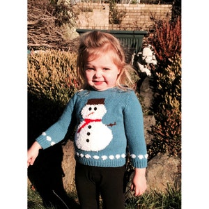 Könnte beinhalten: Ein kleines Kind trägt einen blauen Strickpullover mit einem weißen Schneemann-Motiv. Der Schneemann hat einen roten Schal und schwarze Knöpfe.