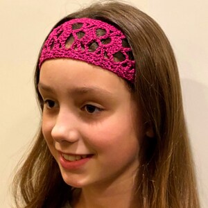 May include: A pink crocheted headband with a delicate floral pattern. The headband is worn by a person with long brown hair.